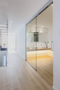 Baño moderno abierto con puerta de cristal, lavabo doble sobre encimera de maderay suelo de madera en vivienda estilo nórdico - Chiralt Arquitectos Valencia